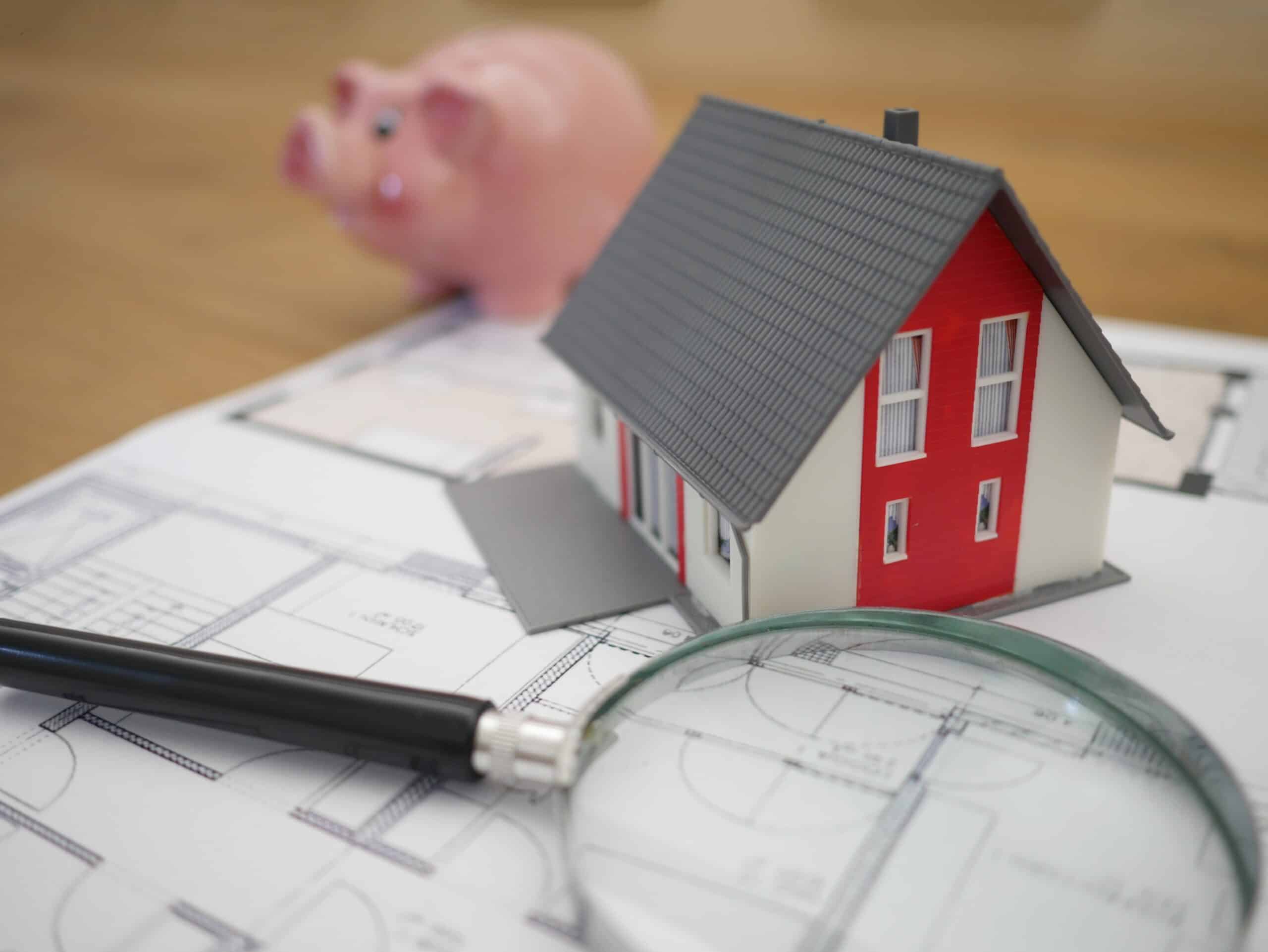 White and red wooden house beside gray framed magnifying glass in front of a piggy bank