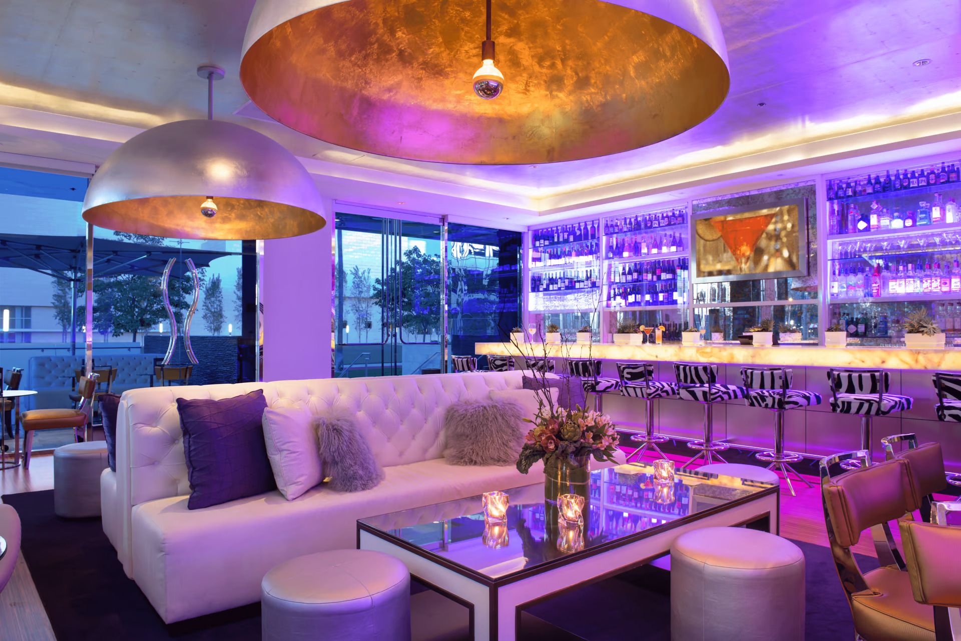 Bar and Lounge Seating Area at Silver Trumpet Restaurant and Bar in Costa Mesa with White Sofa, Glass Top Coffee Table, Zebra print barstools, Large Can Light Chandeliers and Liquor Bottles