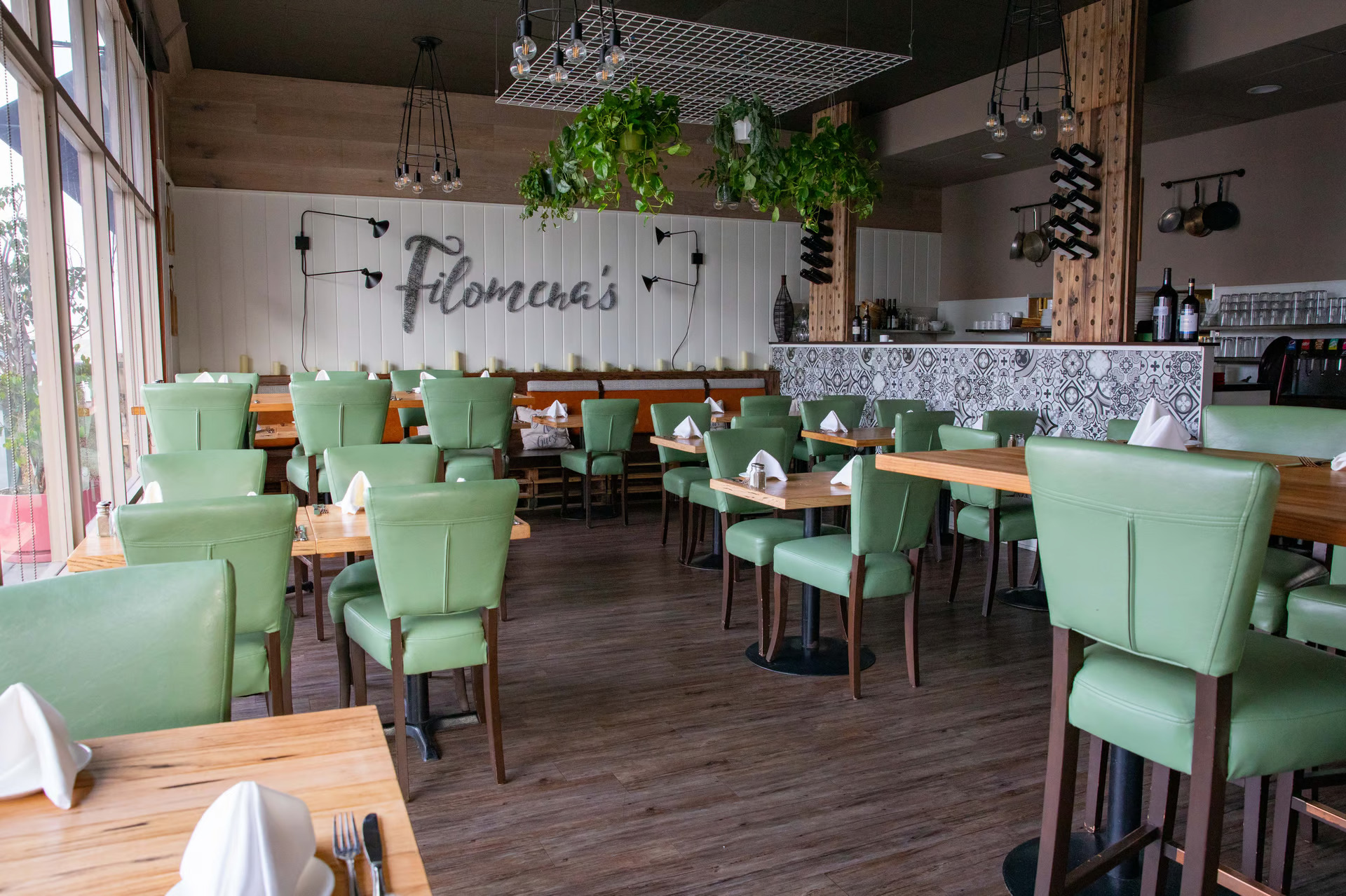 View of Filomena’s Italian Kitchen in Costa Mesa Dining Area with Retro Green Chairs, Folded White Napkins on Wood Tables, and Wine Bottles in Column Holders