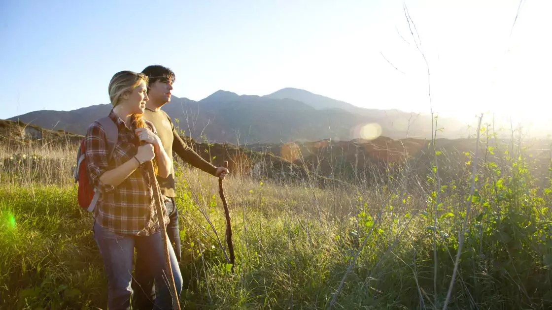 Couple Hiking with Wood Walking Sticks in Tall Grass with Mountains in the Background at O’Neill Regional Park