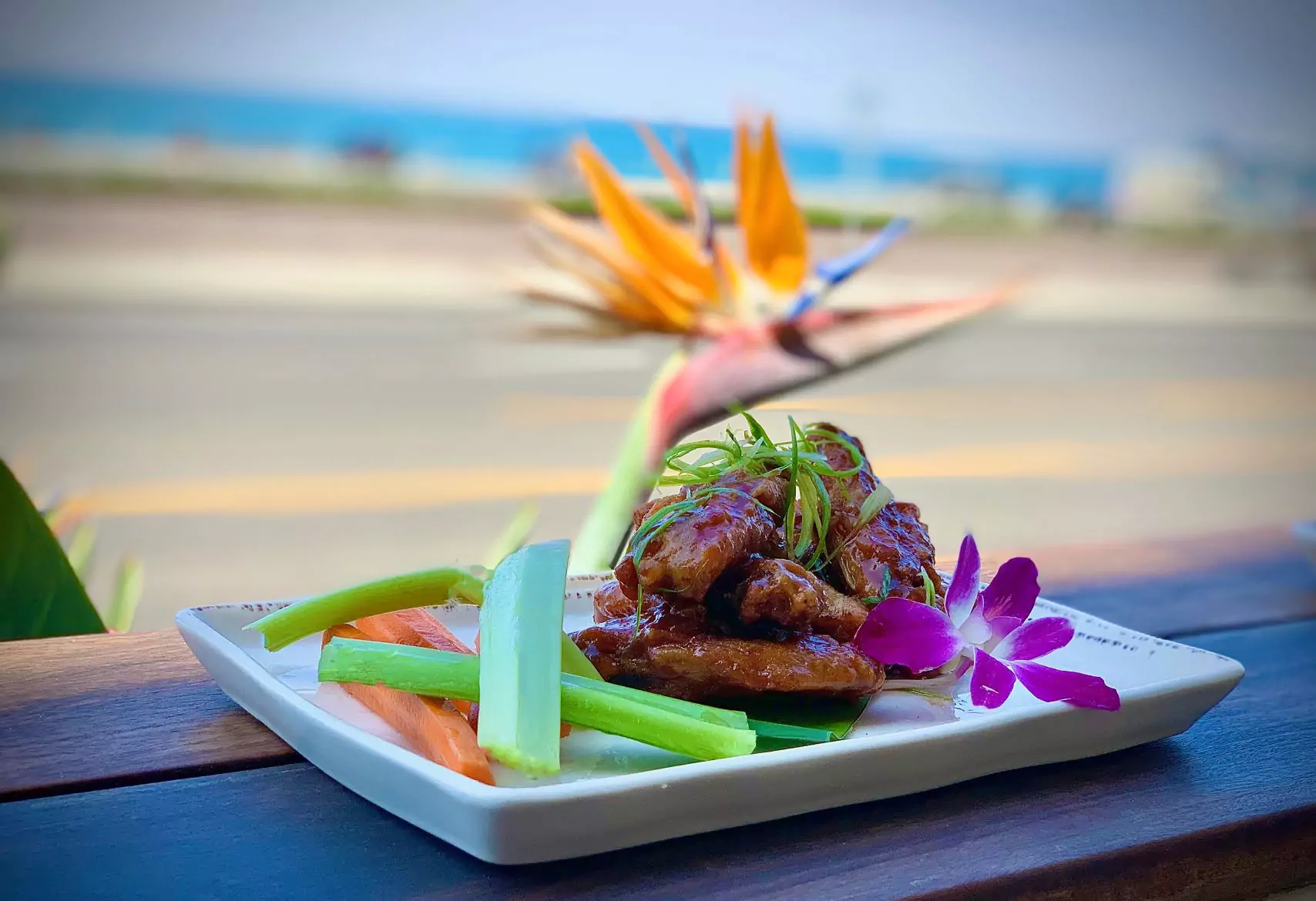 Plate of Wings with Sauce, Garnishes, Celery and Carrot Sticks, and Edible Flowers on Outdoor Patio Dining Table with Views of the Ocean in Background