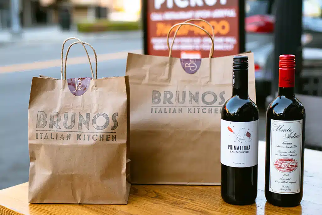 two takeout bags filled with fine italian food and brunos italian kitchen on the front set beside two bottles of red wine