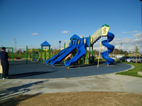 Playground with Blue, green, and yellow sets and walking sidewalks in the foreground at the Fountain Valley Recreation Center & Sports Park