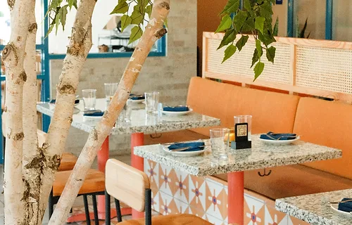 View of Interior of Ini Restaurant with Granite Topped Tables, Multi colored orange and white mosaic tiles, orange suede benches and seat cushions, and a medium sized tree in the foreground