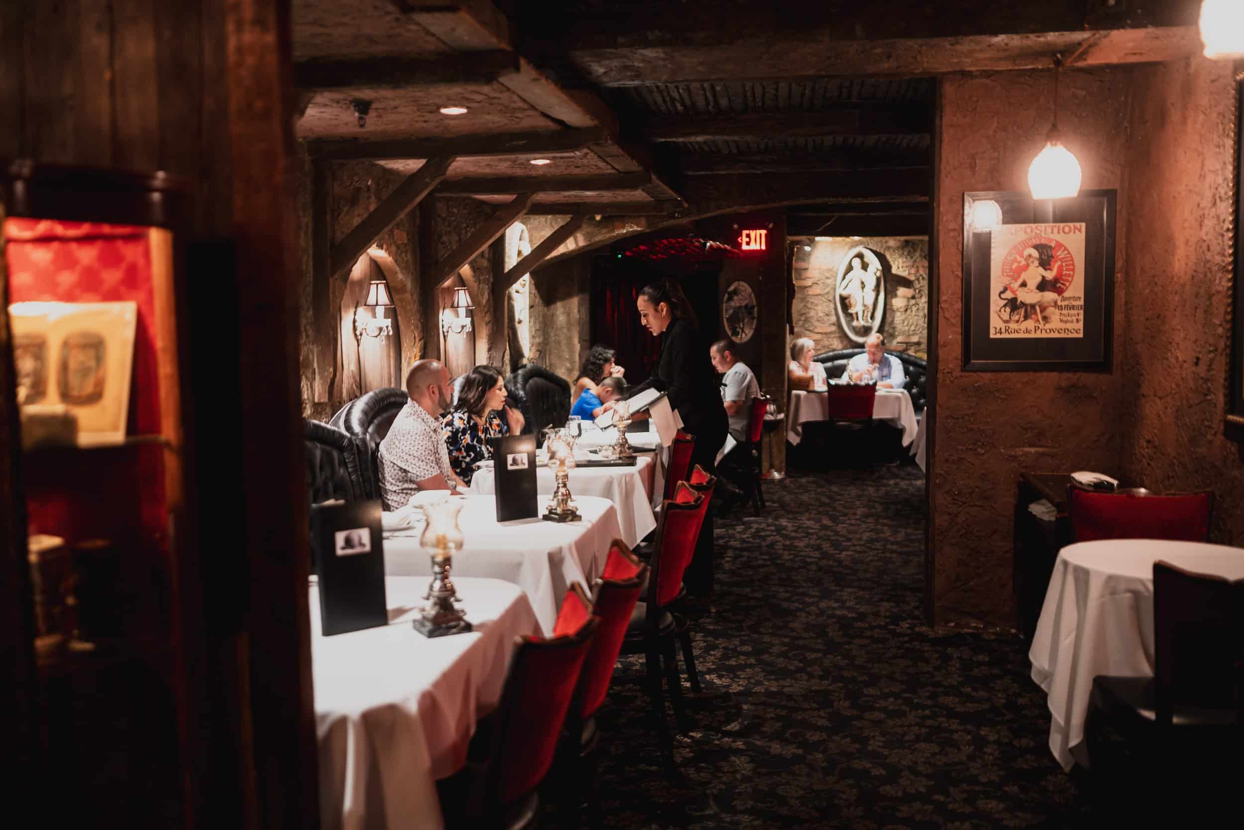 Diners sitting in a dimly lit upscale restaurant with a mostly brown and red themed color scheme and white tablecloths at The Cellar Restaurant