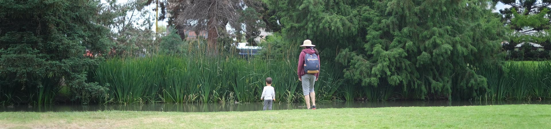 Mom wearing a maroon hoodie, straw brimmed hat, gray capri pants, and backpack with young toddler child looking at a pond and reed plants at the Fullerton Arboretum and Botanical Garden