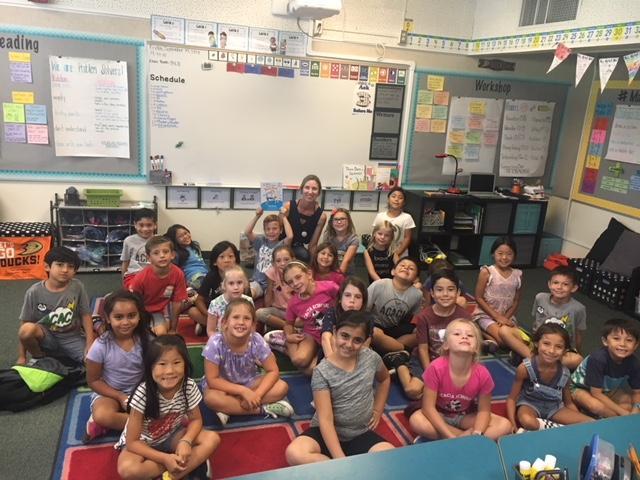 Teacher and students sitting and posing in classroom with chalkboard and dry eraser board with school lessons and notes in background at Acacia Elementary