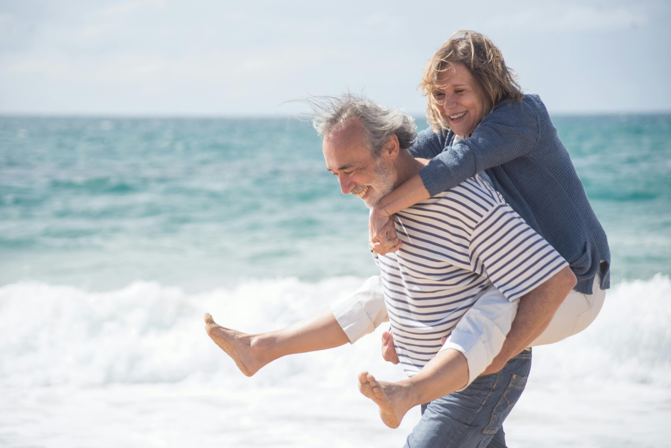 Older couple at the beach, man holding woman as waves crash in the distance, enjoying their new lifestyle after downsizing their Laguna Hills property to a more manageable home
